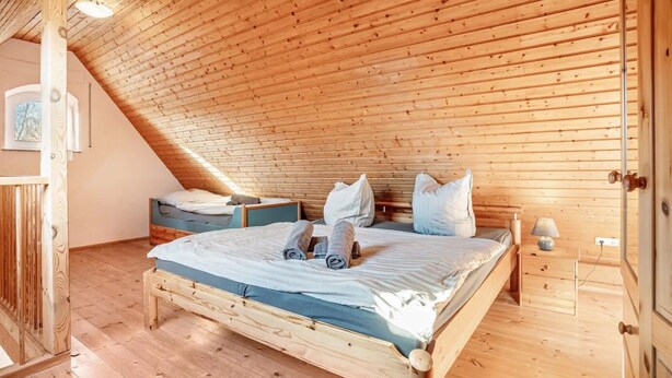 Quarto com teto inclinado, paredes de madeira, cama com almofadas, mesa de cabeceira e abajur.