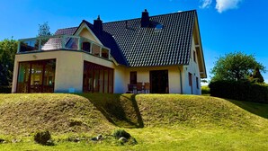 Uma casa de dois andares com uma varanda, uma janela grande e uma colina gramada ao primeiro plano.