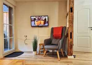 A living room with a television mounted on the wall, a grey armchair with a red throw, a small wooden stool, a potted plant, and a white door.
