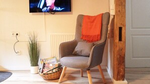 Ein Wohnzimmer mit grauem Sessel, einem an der Wand befestigten Fernseher, einem Blumentopf und einem Korb mit Zeitschriften.