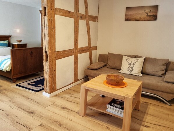 A room with a wooden floor, a bed with a blue and white striped bedspread, a wooden dresser, a brown sofa with a deer pillow, and a small wooden table with a bowl.