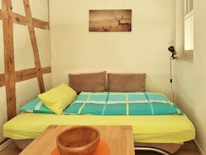 A small, cozy room with a bed, pillows, a wooden table with a bowl, a picture on the wall, and a window with blinds.