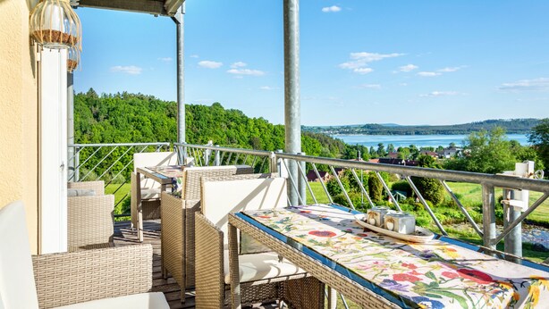 A balcony with wicker furniture, a table with a floral tablecloth, and a view of a lake and forest.