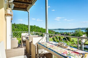 A balcony with wicker furniture, a table with a floral tablecloth, and a view of a lake and forest. - Suite 'Seelixir' with Lake View and Sunny Balcony on a Panoramic Plateau (Spalt / Enderndorf am See)