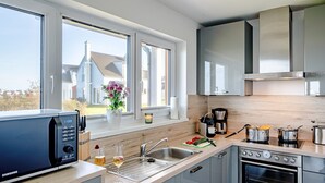 A modern kitchen with a built-in microwave, stainless steel appliances, a large window, and a view of a house. - Holiday Home 'Am Meer' with Sea View, Private Terrace and Wi-Fi (Kappeln)