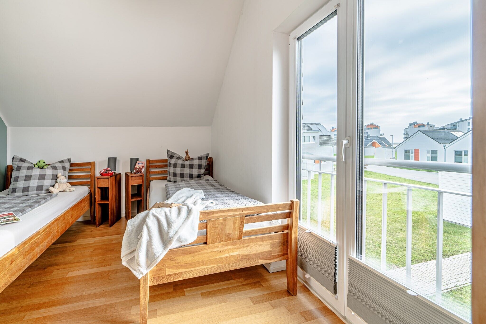 A bedroom with two wooden beds, a window with a view of houses, and a small table with a teddy bear.
