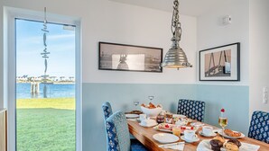 A dining area with a view of the sea, a wooden table set with plates, glasses, and pastries, and patterned blue chairs. - Holiday Home 'Am Meer' with Sea View, Private Terrace and Wi-Fi (Kappeln)