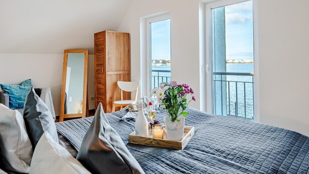 A bedroom with a bed, a bedside table with a tray, a mirror, and a view of the water. - Holiday Home 'Am Meer' with Sea View, Private Terrace and Wi-Fi (Kappeln)