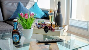Une table basse en verre sur laquelle on trouve une bouteille de bière, une télécommande, un livre, un verre de champagne, un bouquet de tulipes et une boîte de chocolats.