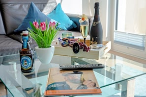 Una mesa de centro de vidrio con una botella de cerveza, un control remoto, un libro, una copa de champán, un ramo de tulipanes y una caja de bombones.