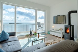 A living room with a sofa, a glass coffee table, a fireplace, a TV, and a balcony with a view of the sea.