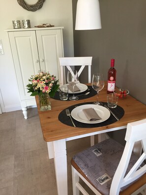 Ein Esstisch ist gedeckt mit Tellern, Gläsern und einer Flasche. Im Hintergrund steht eine Blumenvase und eine Lampe.
