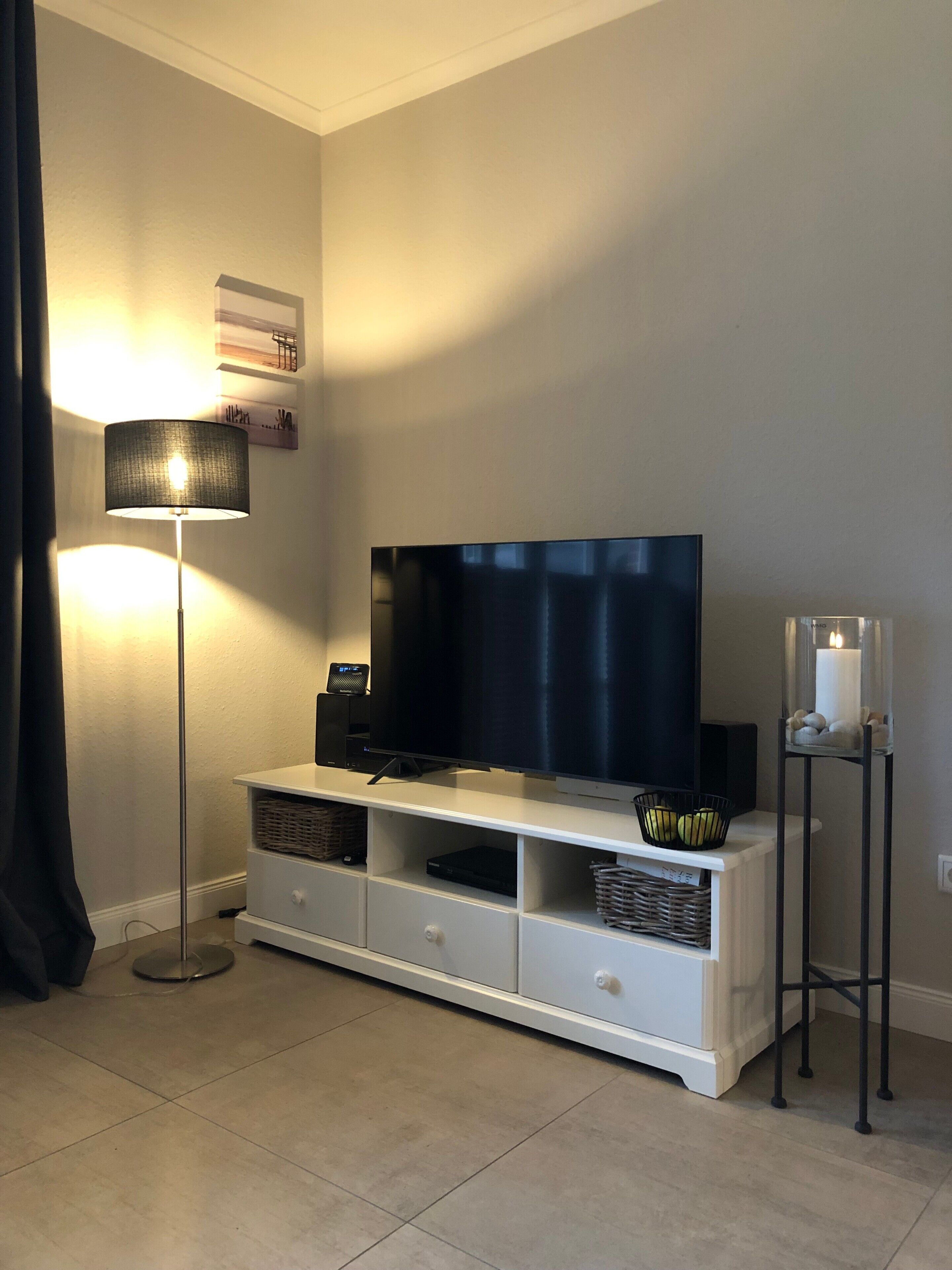A living room with a flat-screen TV on a white entertainment unit, a floor lamp, and a candle holder.