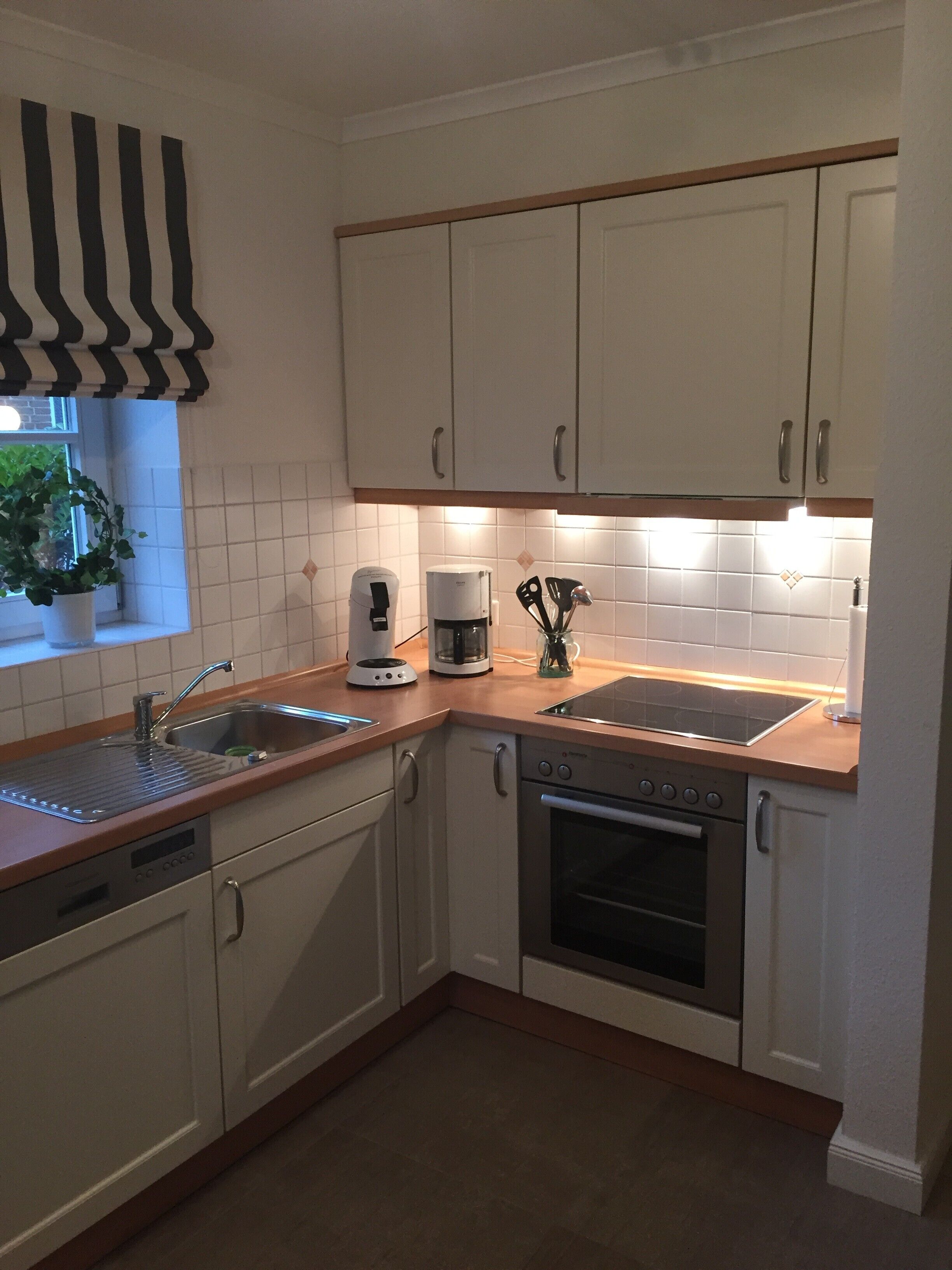 Eine Küche mit weißen Schränken, einem Geschirrspüler, einem Backofen, einer Kaffeemaschine und einem Fenster mit einer Pflanze.