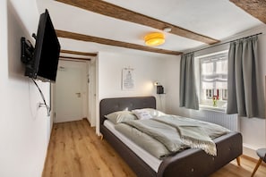A bedroom with a bed, a mounted TV, a ceiling light, a window with curtains, and a door.