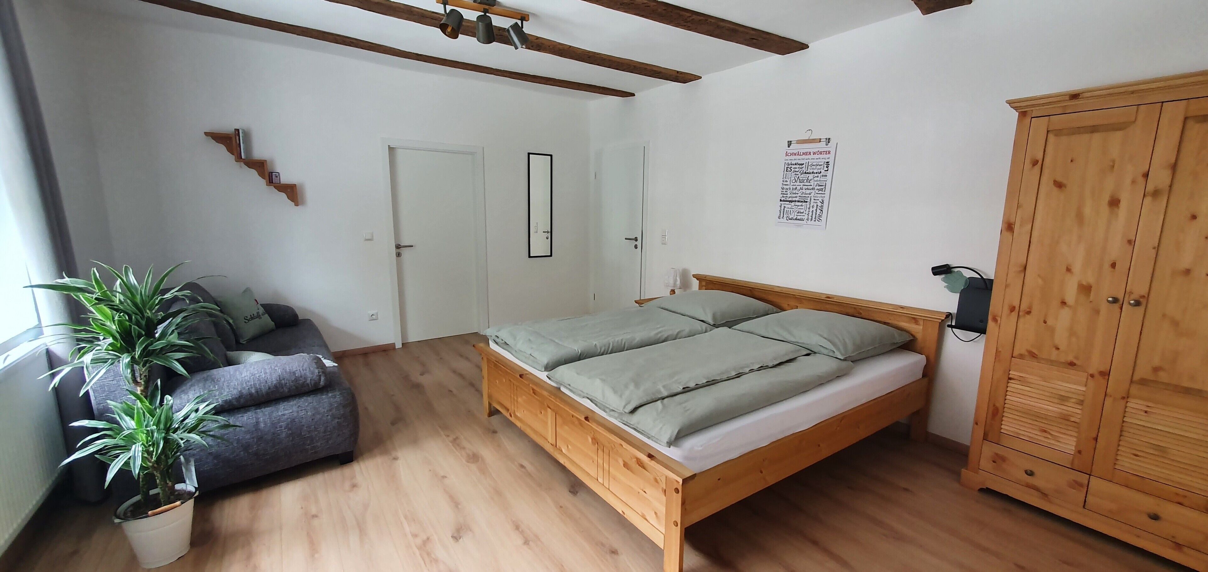 Een slaapkamer met een houten bed, een grijze bank, een kamerplant, een kledingkast en een deur.