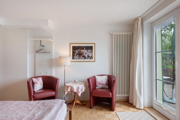 Habitación de hotel con dos sillones rojos, una mesita con lámpara, un cuadro enmarcado en la pared y un ventanal con cortinas.