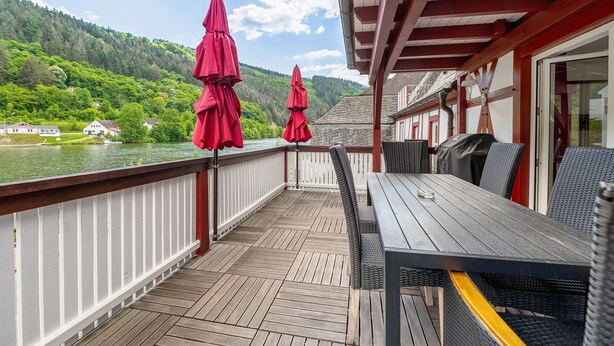 A wooden deck with a table and chairs, overlooking a lake and forested hills. - Holiday Home 'Moseljuwel' with Sauna, Pool & Large Moselle Terrace (Traben-Trarbach)