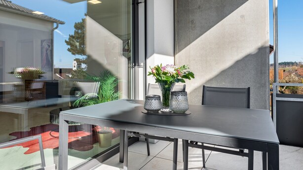 Un espace de restauration extérieur moderne, doté d’une paroi en verre, d’une table agrémentée de deux vases et de chaises.