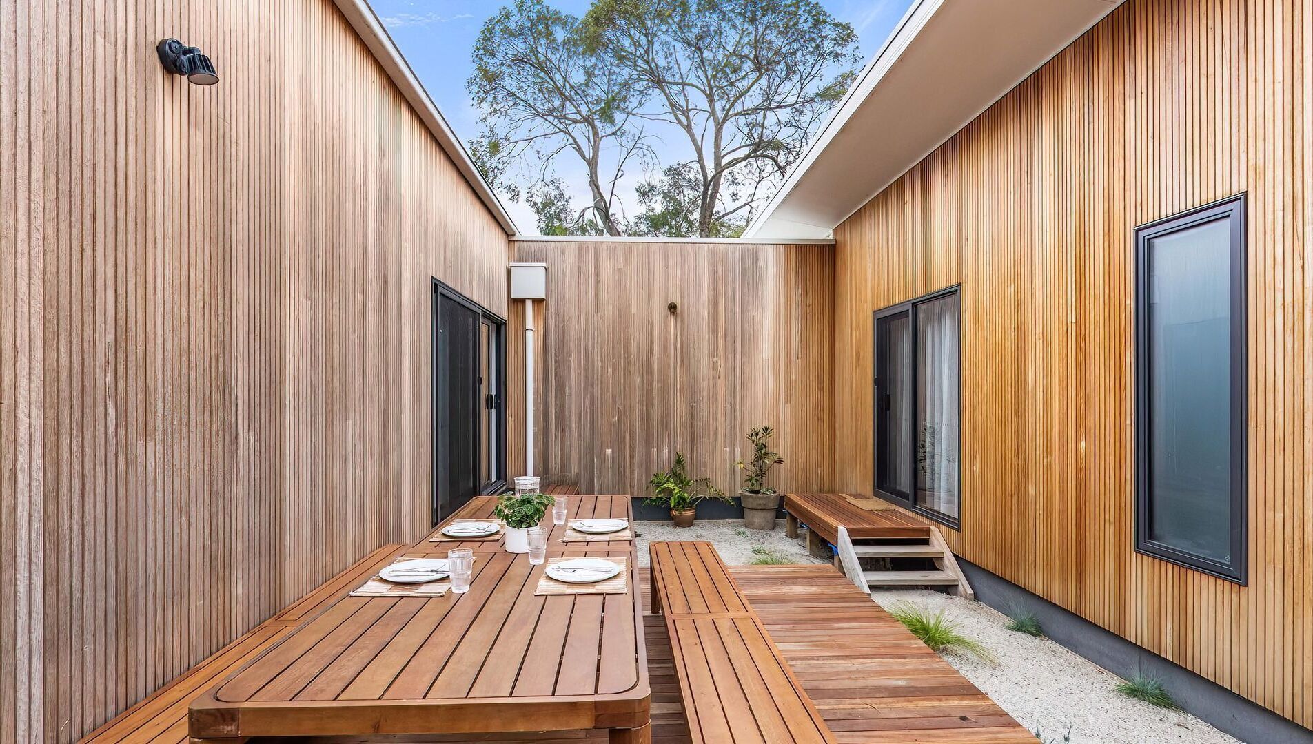 A modern outdoor dining area with wooden tables and benches, surrounded by vertical wooden panels and large windows.