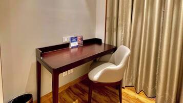 A room with a wooden desk, a white chair, a small trash bin, and a window with curtains.