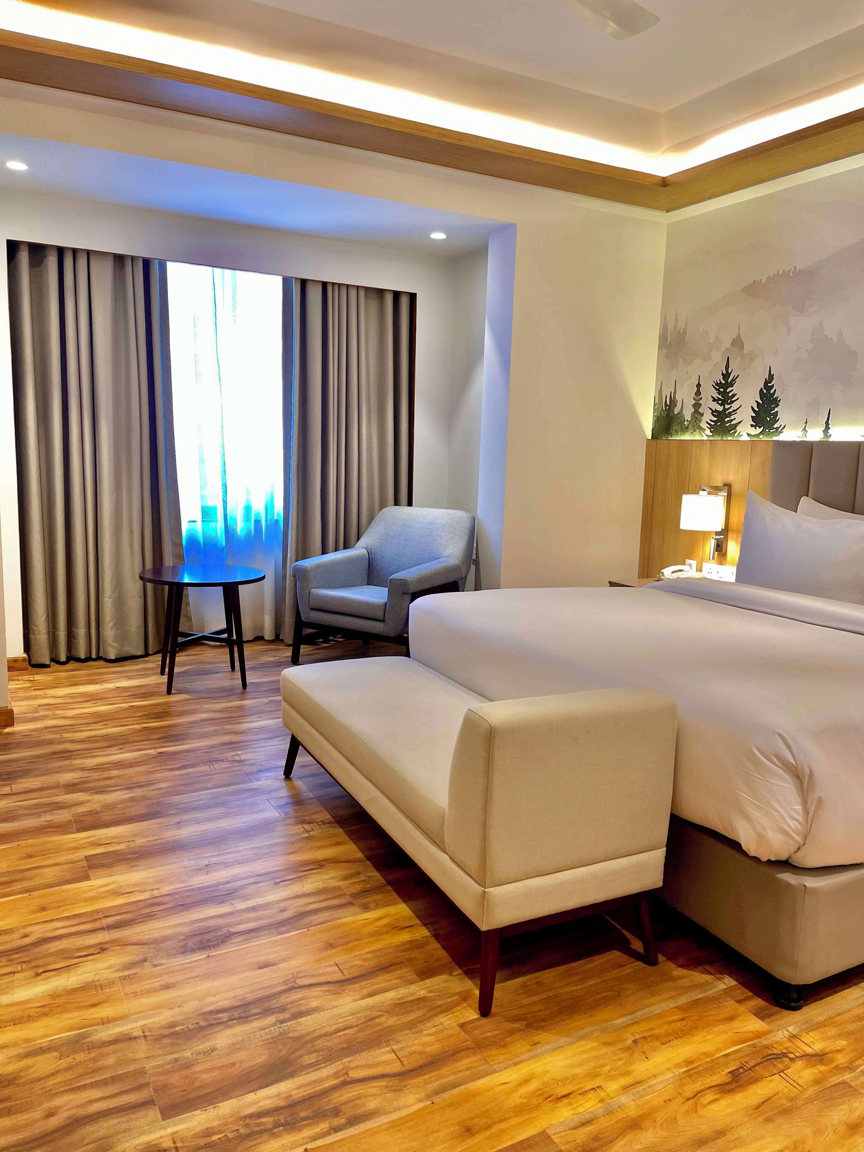 Une chambre d’hôtel avec un grand lit, un canapé, un fauteuil, une petite table et une fenêtre avec des rideaux.