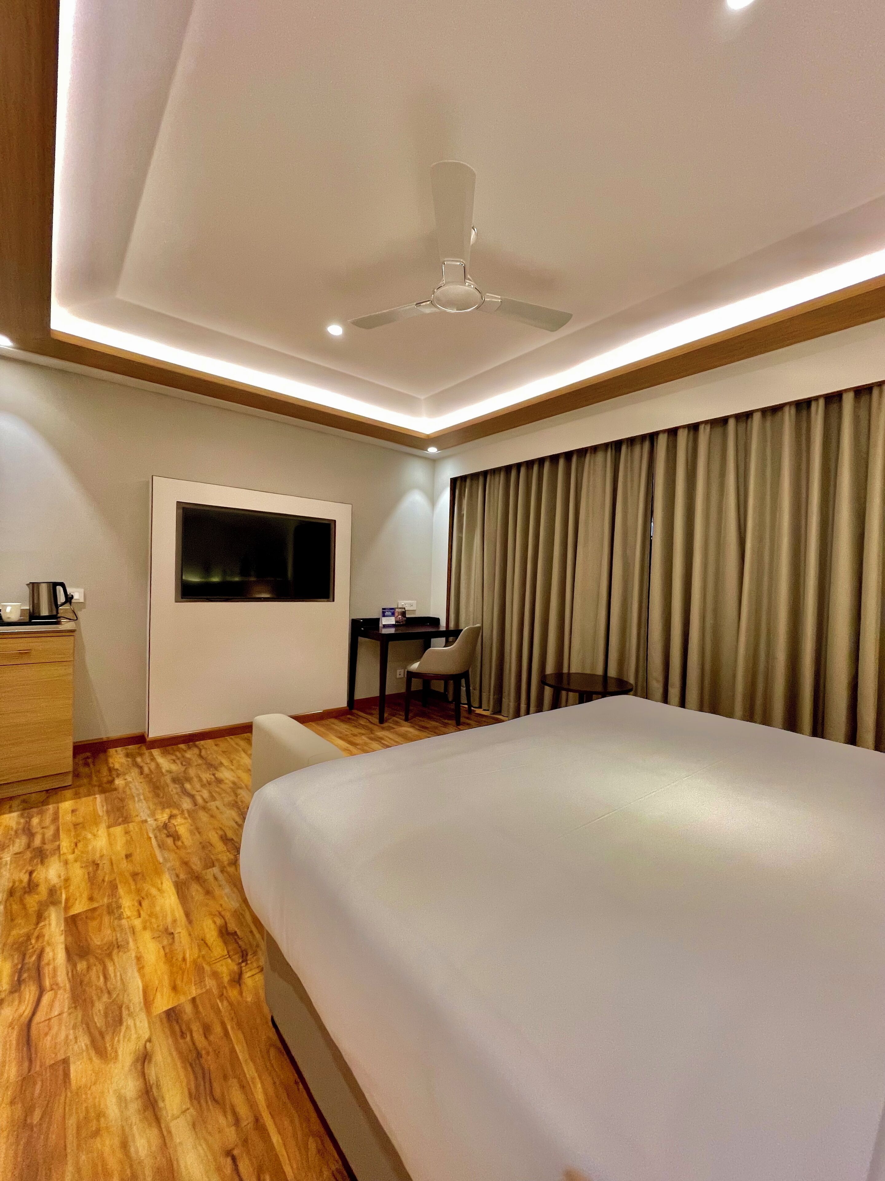 A hotel room with a large bed, a desk with a chair, a TV mounted on the wall, and a ceiling fan.
