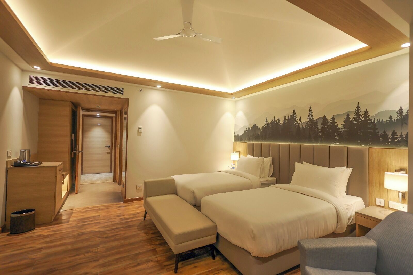A hotel room with two beds, a desk, and a chair. There is a ceiling fan, a wall-mounted TV, and a painting of a forest on the wall.