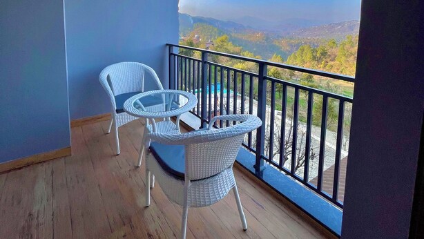 Un balcon agrémenté de deux fauteuils en osier et d’une petite table, offrant une vue imprenable sur un paysage magnifique, avec des montagnes et un ciel d’un bleu éclatant.