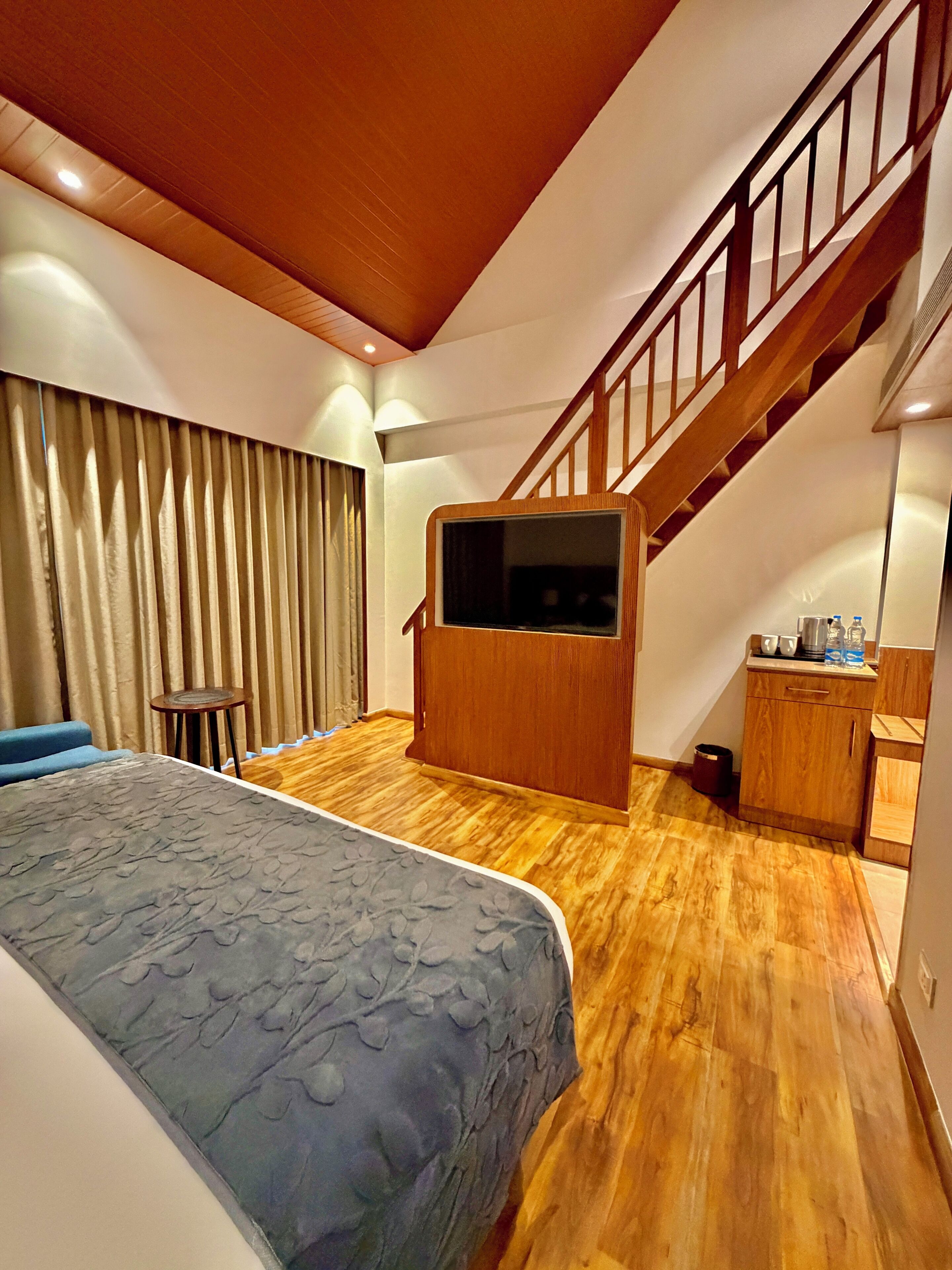 A bedroom with a bed, a wooden staircase, a small table, and a kitchenette area.