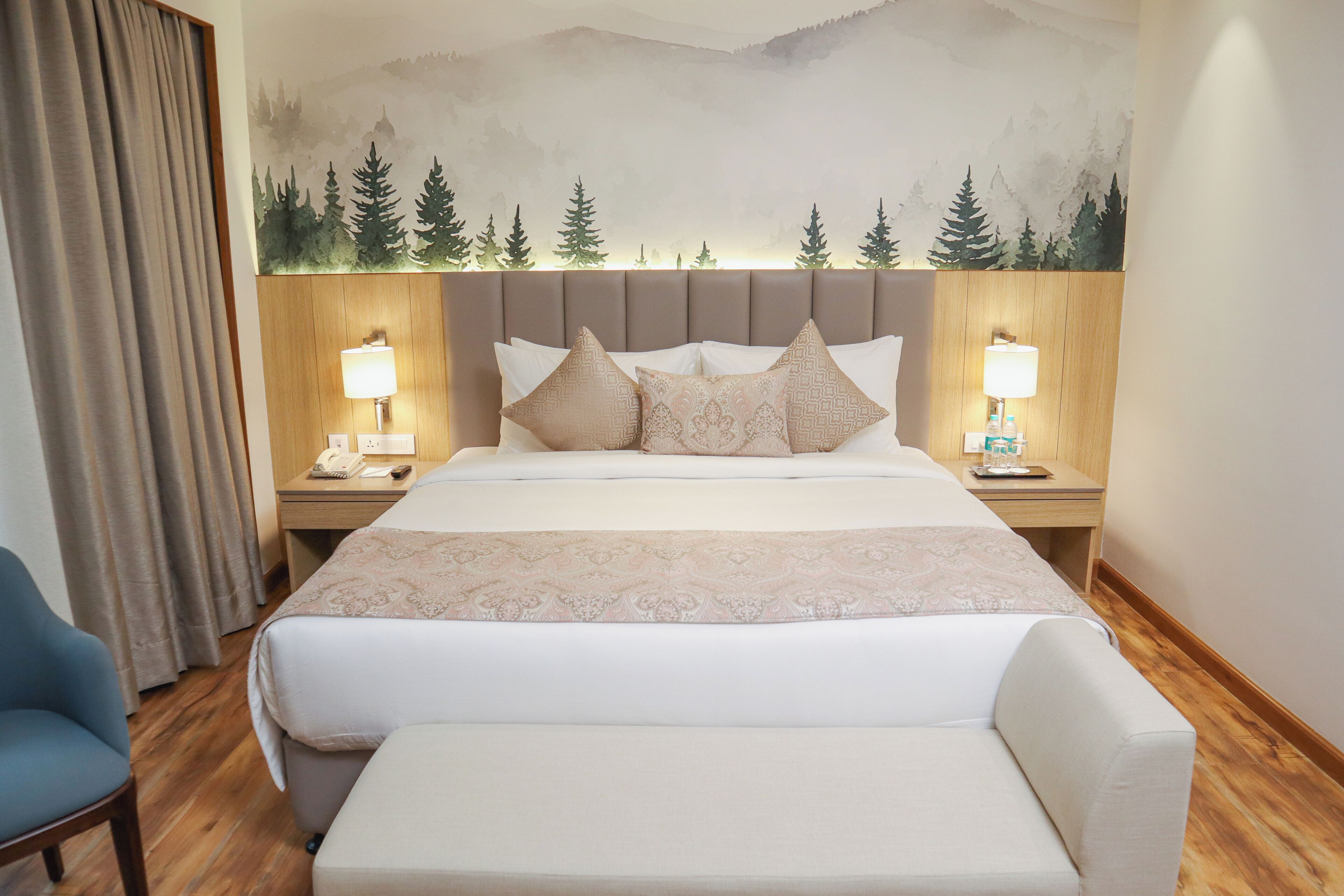 Habitación de hotel con una cama grande, dos mesitas de noche con lámparas, un cabecero de madera, un sillón azul y un mural con árboles en la pared.