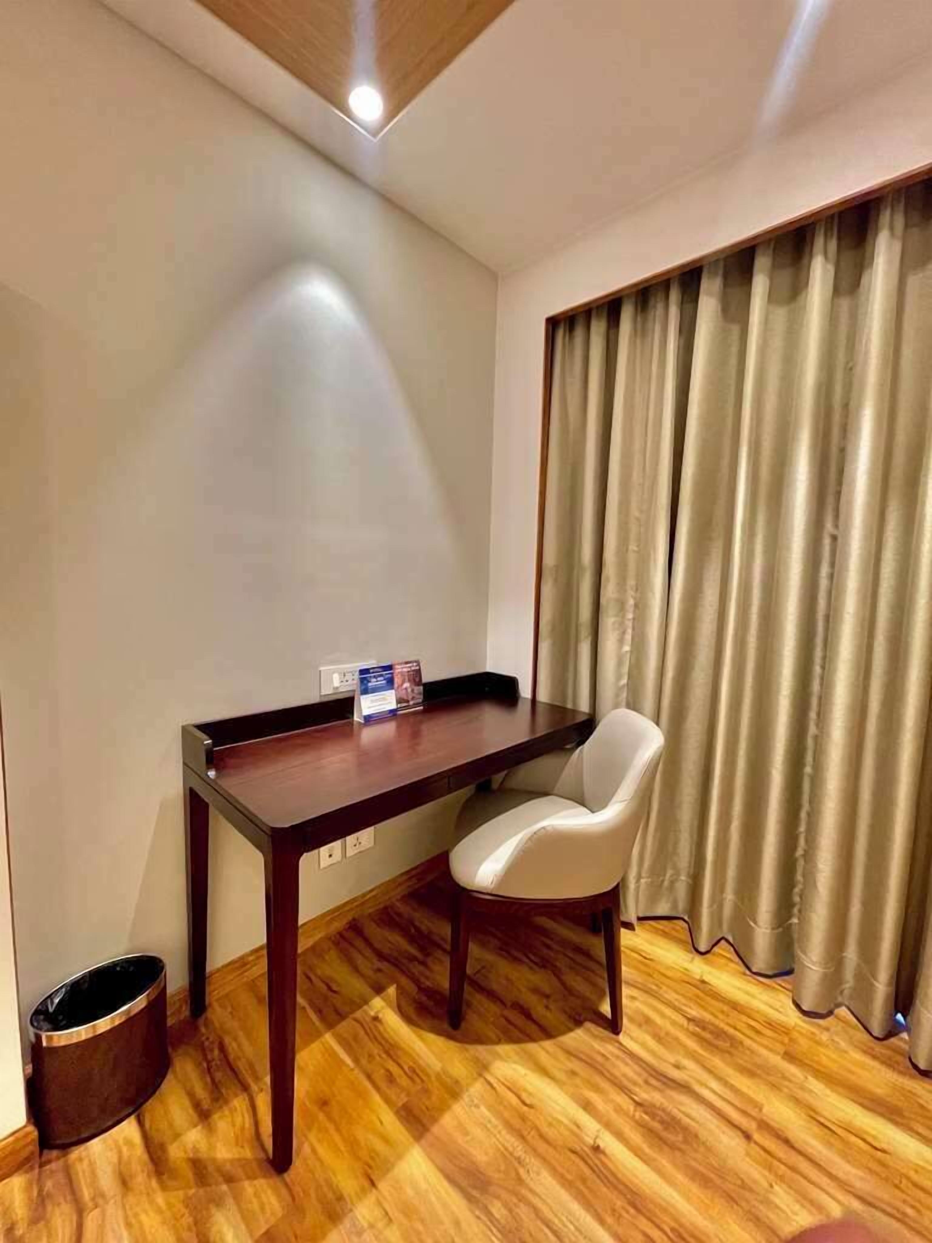 A small room with a wooden desk, a white chair, a trash bin, and a window with curtains.