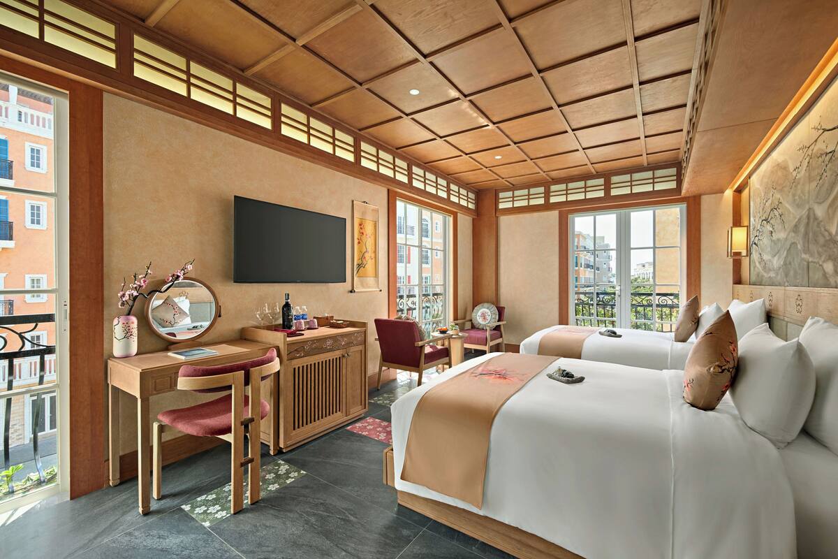 A hotel room with a wooden desk, two beds, a large window, and a view of buildings.