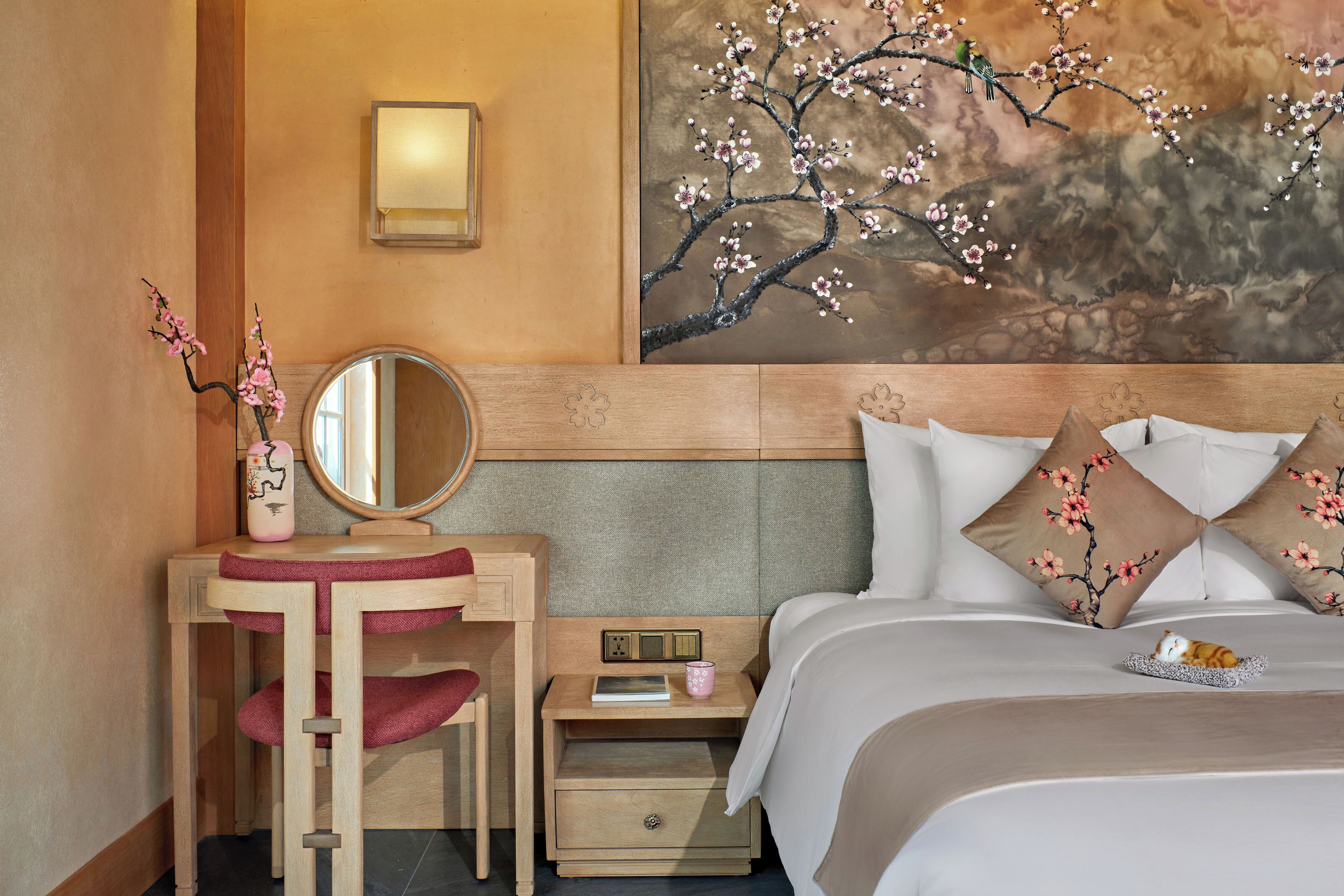 A hotel room with a wooden headboard, a bedside table with a lamp, a mirror, a small nightstand with a book and a pink mug, and a bed with floral pillows.