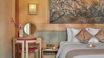 A hotel room with a wooden headboard, a bedside table with a lamp, a mirror, a small nightstand with a book and a pink mug, and a bed with floral pillows.