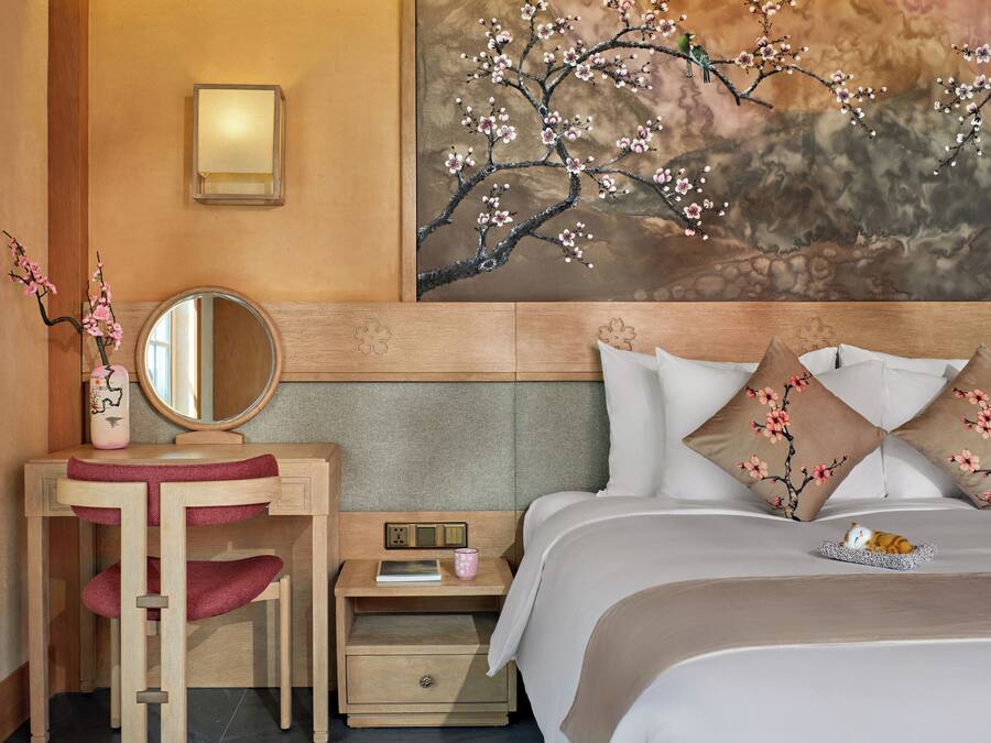 A hotel room with a wooden headboard, a bedside table with a lamp, a mirror, a small nightstand with a book and a pink mug, and a bed with floral pillows.