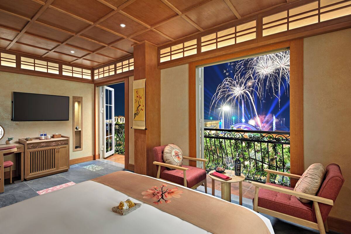 A hotel room with a large bed, a TV, a balcony with a view of fireworks, and a dining area with a table and chairs.