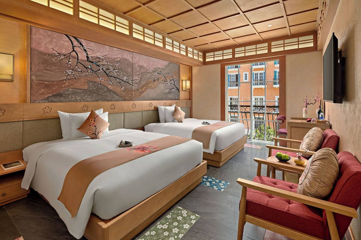 A hotel room with two beds, a sofa, a small table, and a large window with a view of buildings.