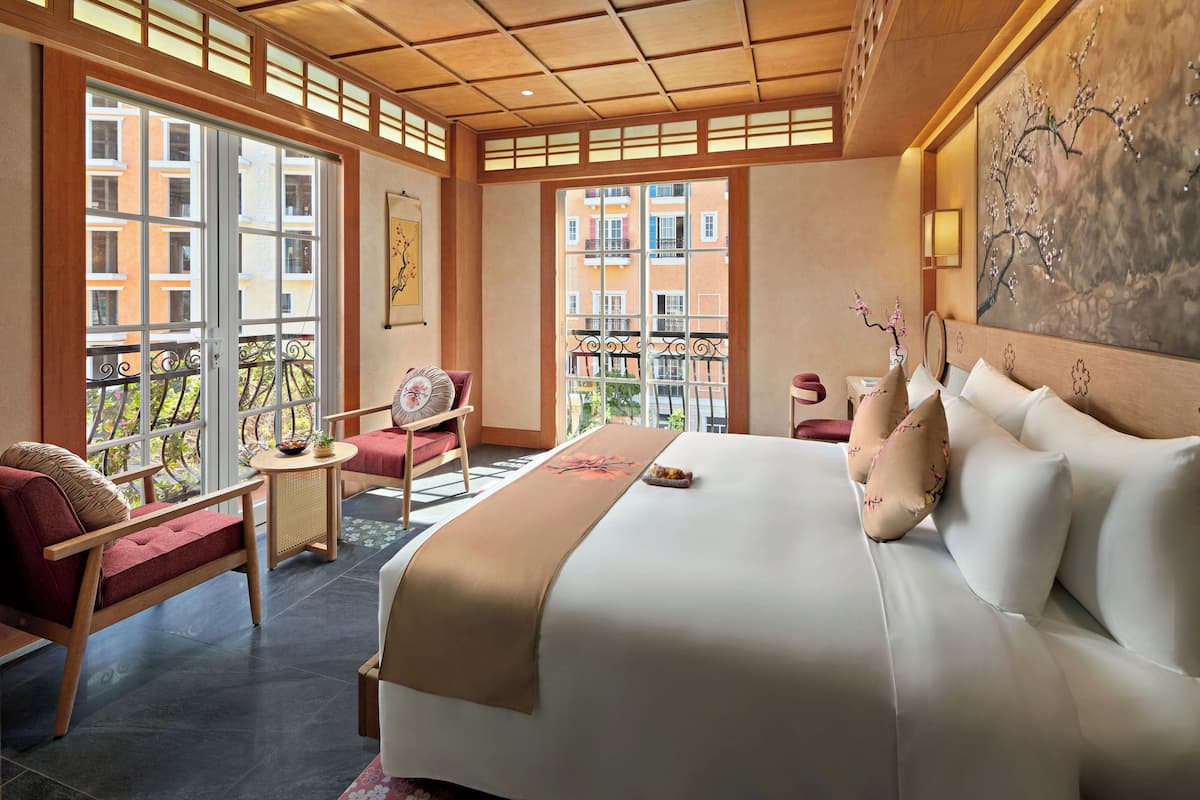 A hotel room with a large bed, a sitting area with a chair and a small table, and a balcony with a view of the city.