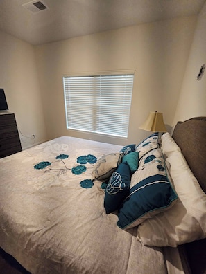 Une chambre à coucher avec un lit, une fenêtre avec des stores, une lampe et une commode.