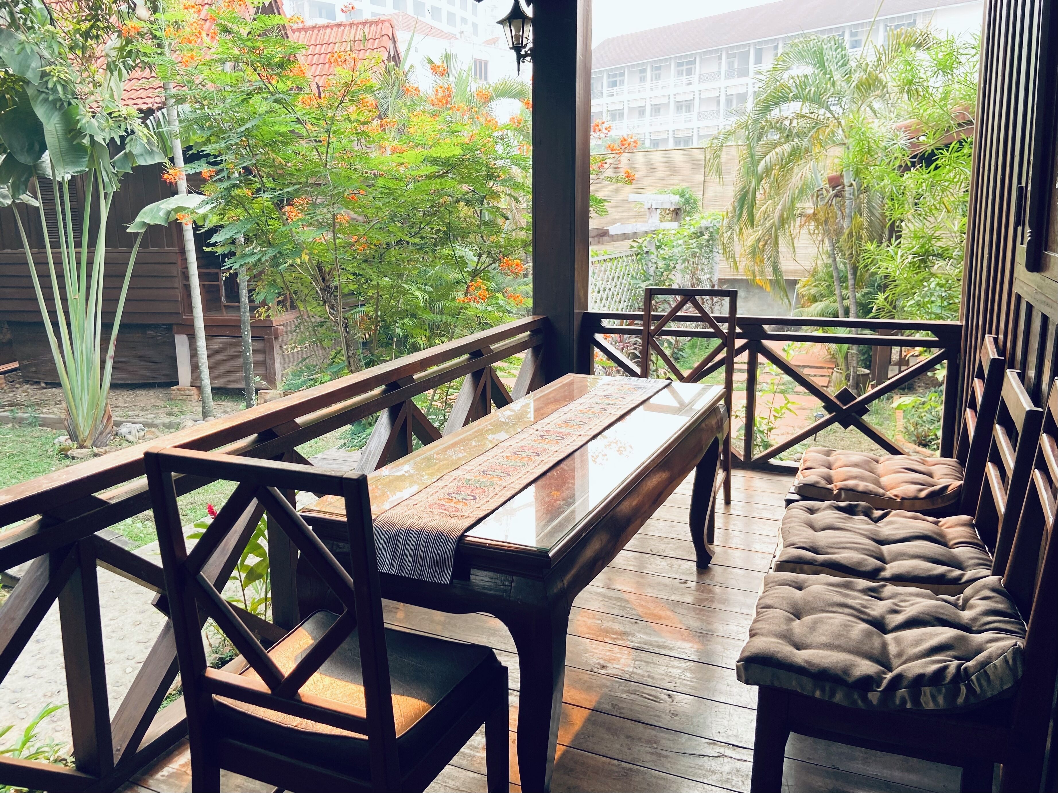 A wooden deck with a bench and a table, surrounded by lush greenery and a view of buildings in the background.