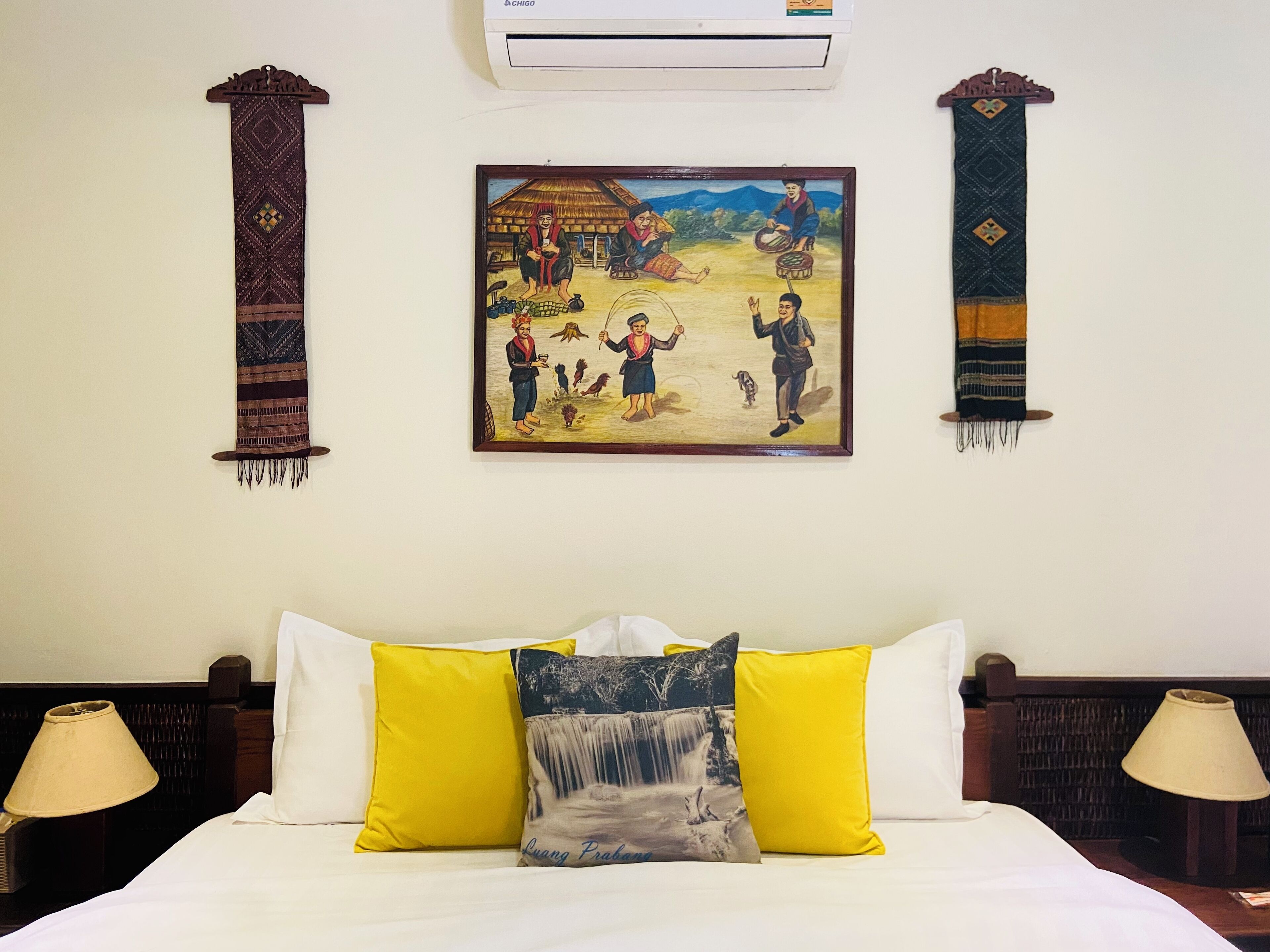 A hotel room with a bed, two nightstands, a wall-mounted air conditioner, and a framed painting depicting a rural scene.