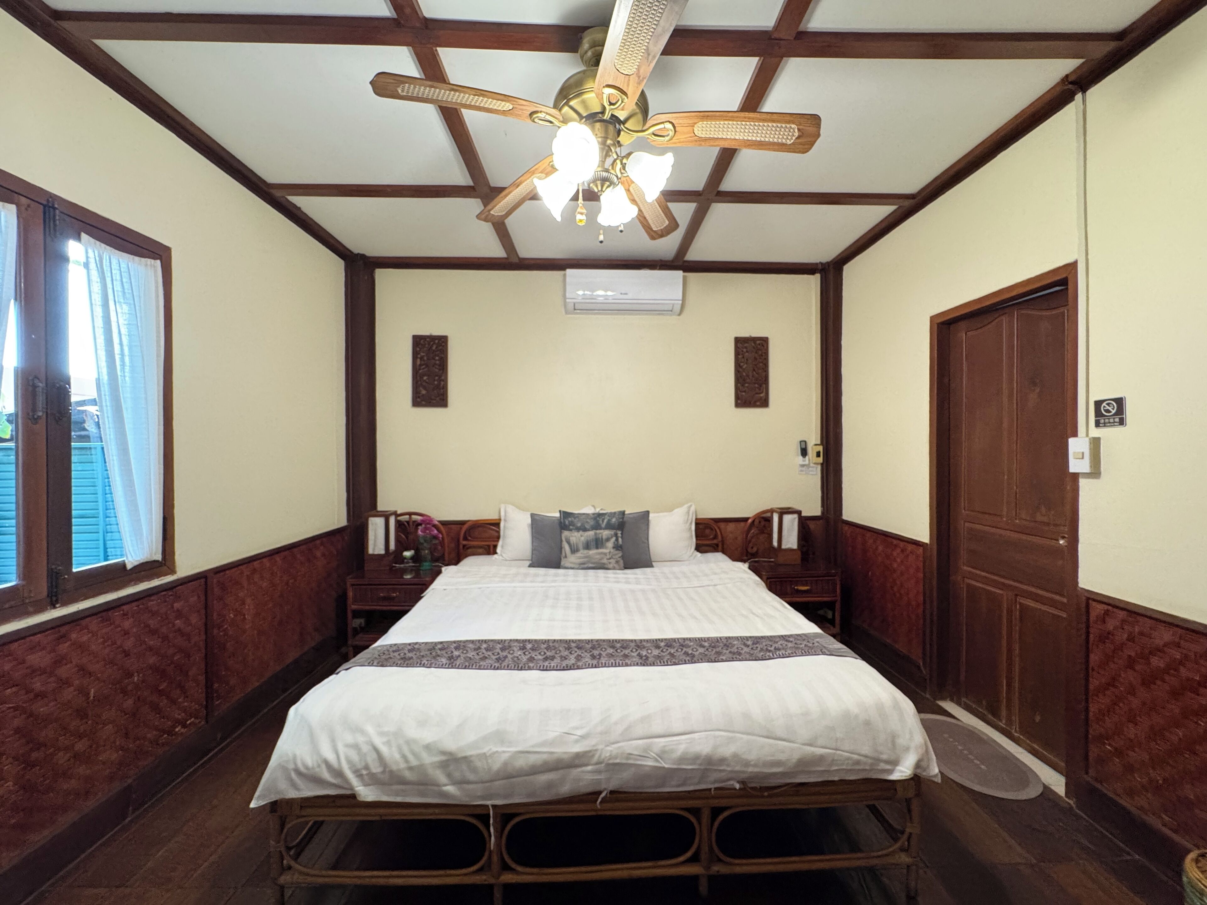 Quarto com cama, cabeceira de madeira, ventilador de teto, janela com cortinas e porta.