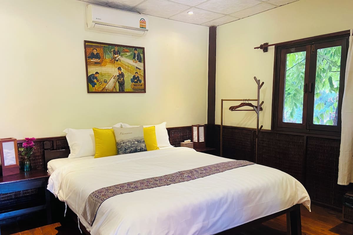 Quarto de hotel com uma cama grande, um quadro na parede, uma janela com vista para vegetação e piso de madeira.