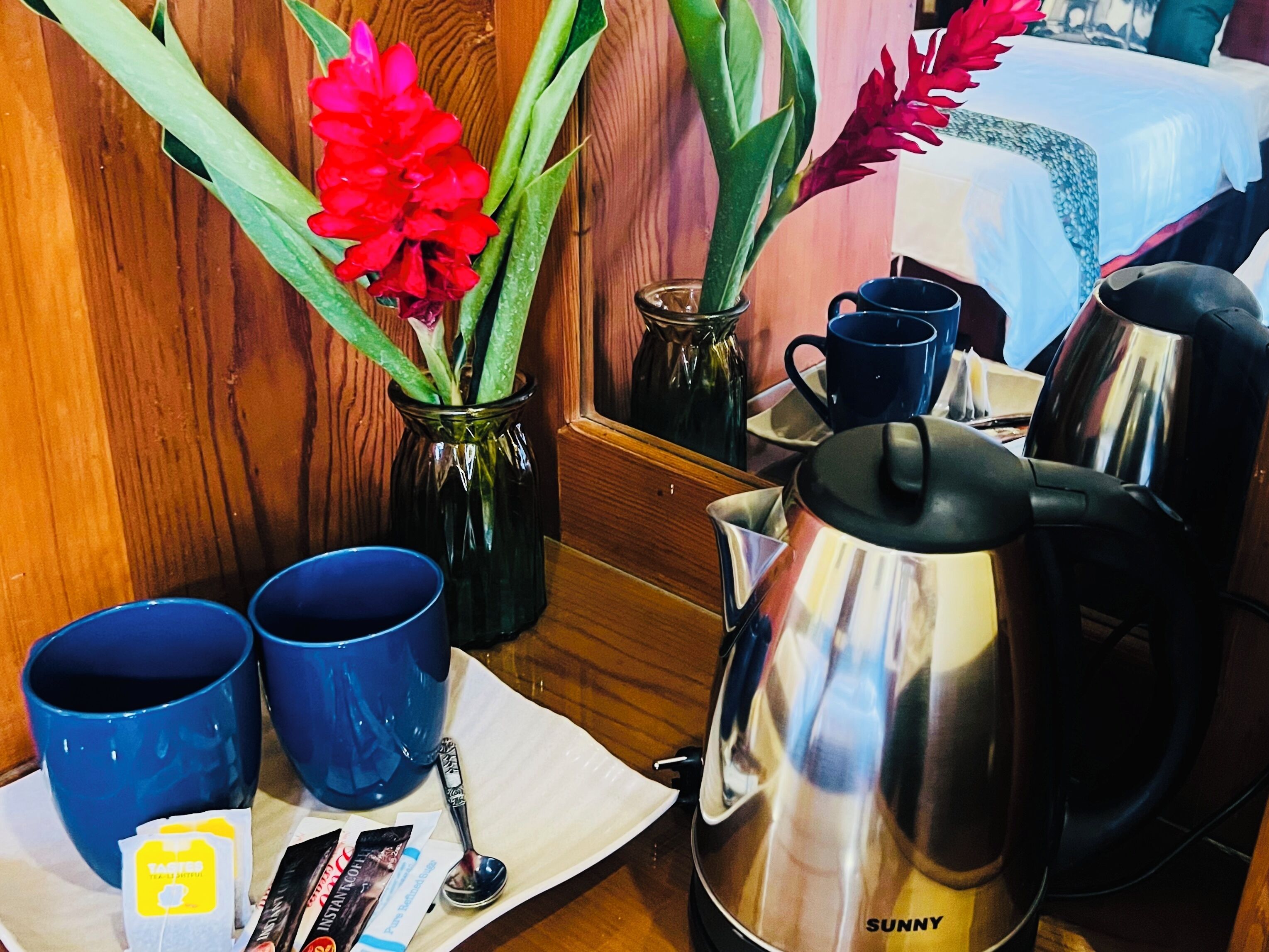 A wooden table with a coffee pot, blue cups, a spoon, and a vase with red flowers.