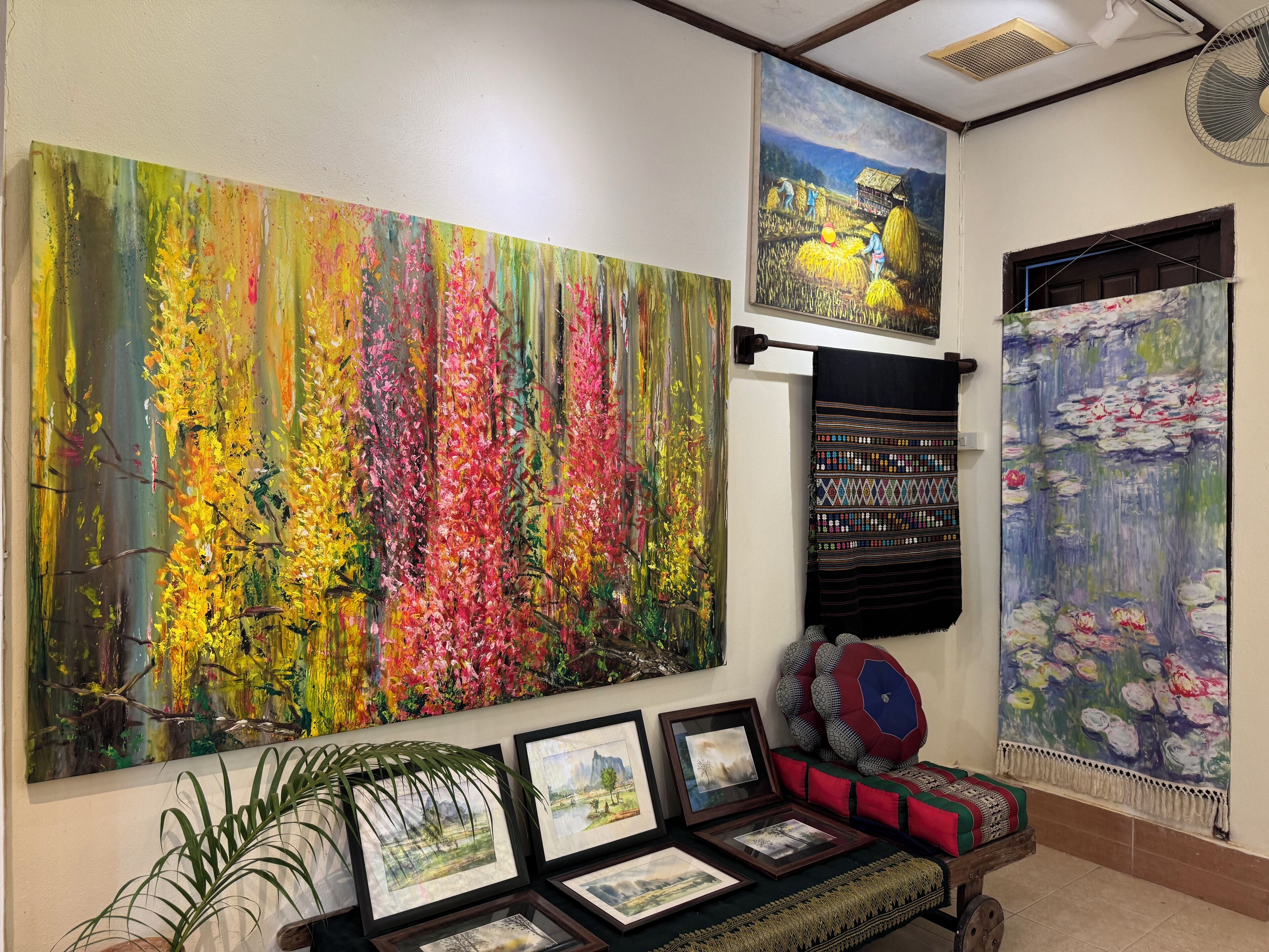 A room with a large colorful painting, a bed with patterned bedding, and a small table with framed pictures.