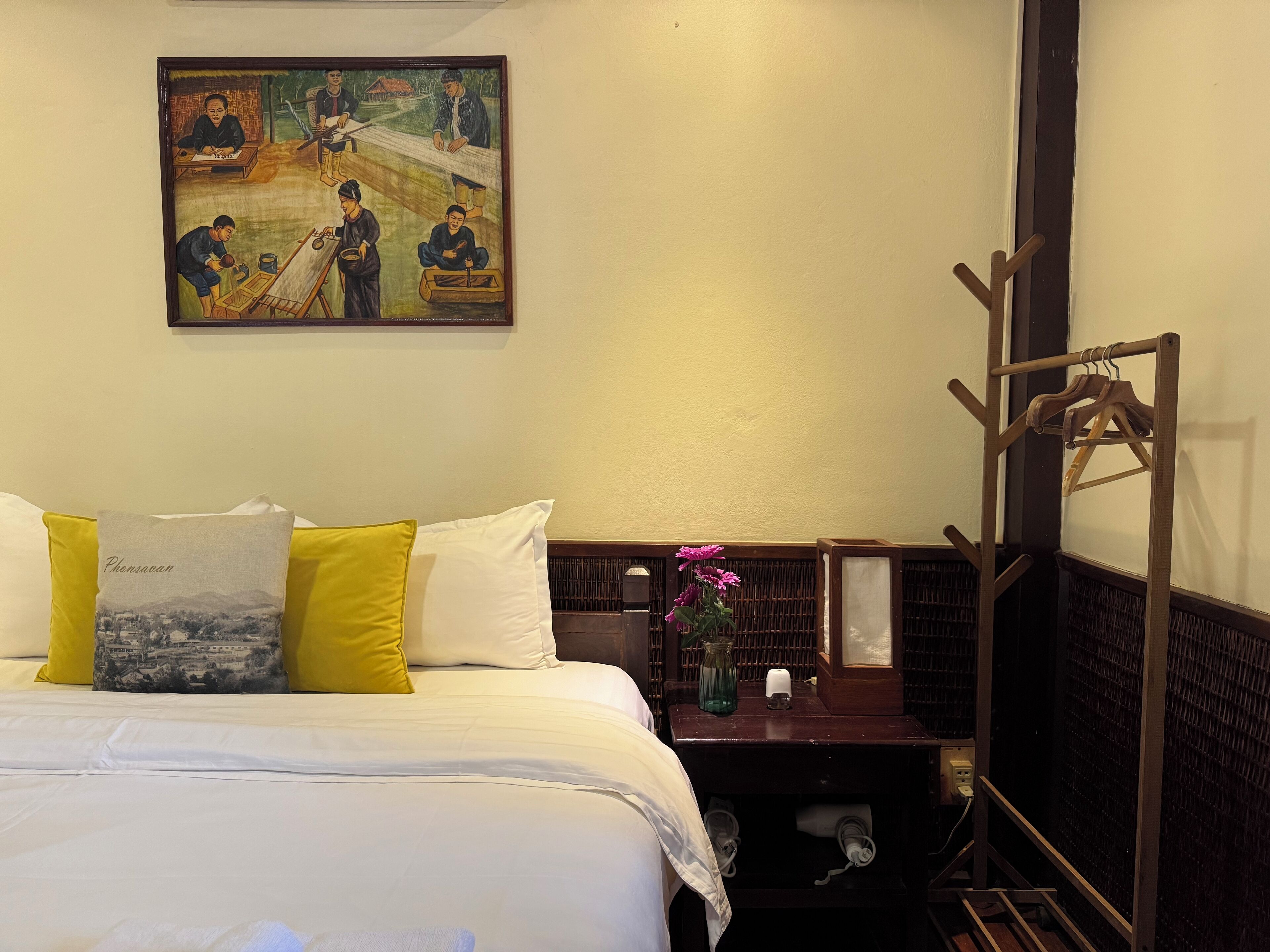 Habitación de hotel con cama, almohadas, un cuadro, un perchero de madera y una pequeña mesa con un florero.