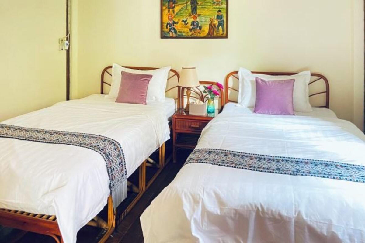 Two single beds with white linens and decorative pillows, a nightstand with a lamp, and a painting on the wall.