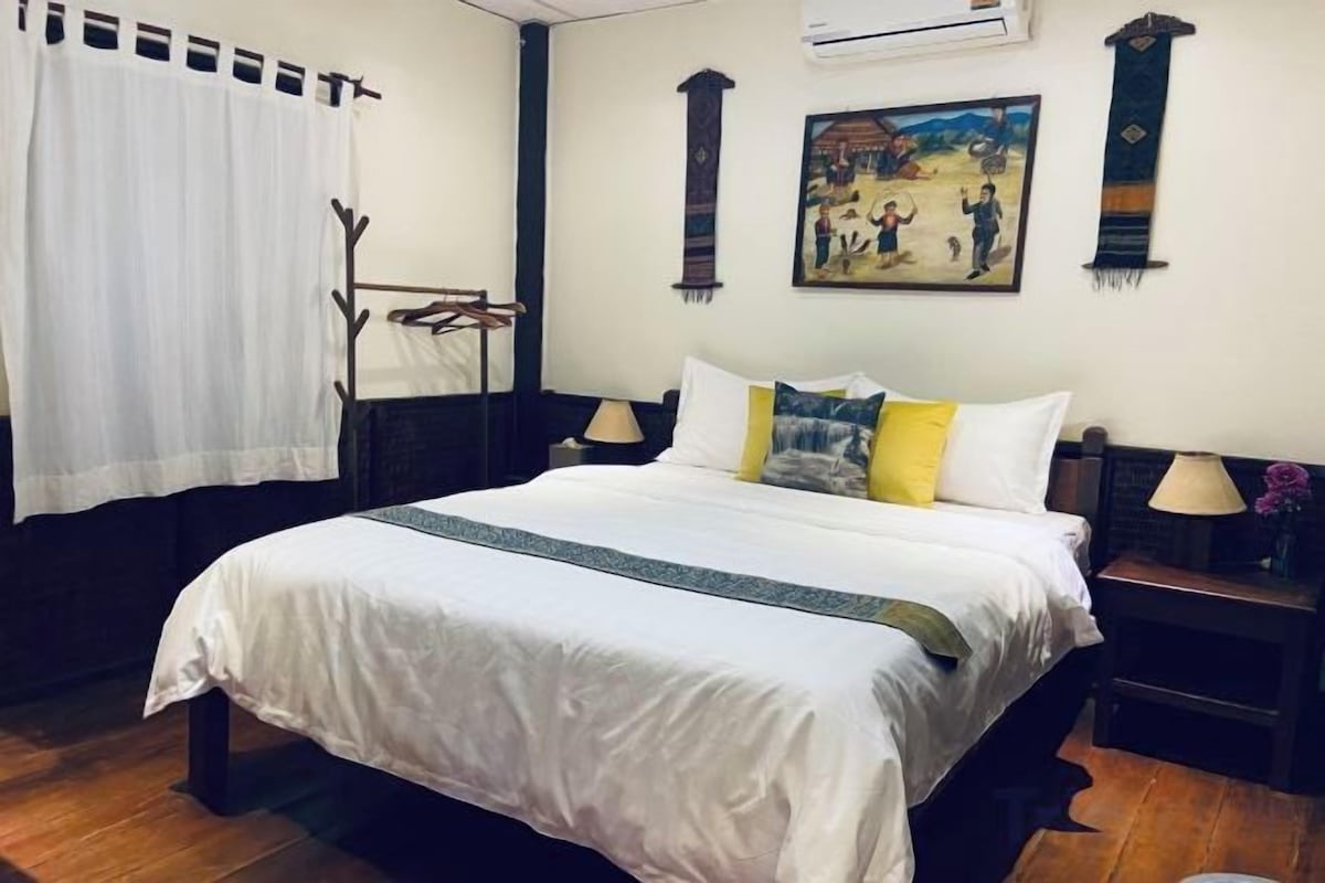 A hotel room with a large bed, a bedside table with a lamp, a wall-mounted air conditioner, and a painting above the bed.