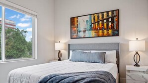 Quarto de hotel moderno com uma cama grande, duas luminárias de cabeceira, uma pintura de paisagem urbana e uma janela com vista para árvores e edifícios.